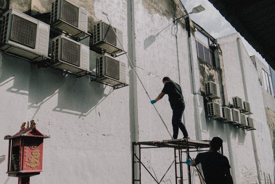 Photo by Putra Rangga on Pexels Two workers on scaffold maintain air conditioning units attached to a building, displaying urban upkeep.