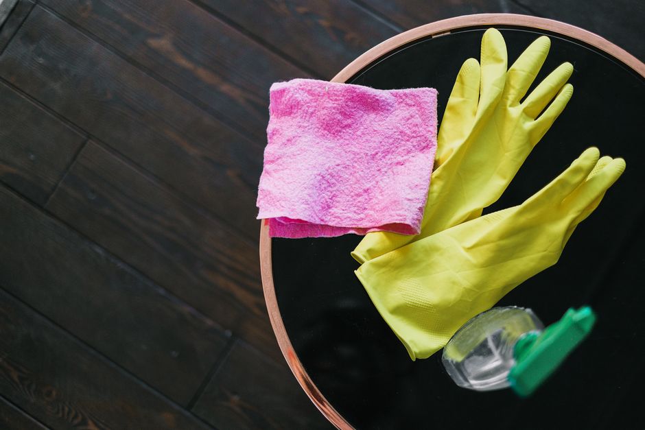 From above of yellow latex gloves placed near rag and spray detergent on table against wooden floor in daytime