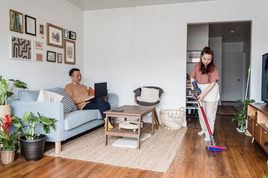 Woman cleaning the wooden floor of a bright living room while a man relaxes on the sofa. Photo by Annushka  Ahuja on Pexels
