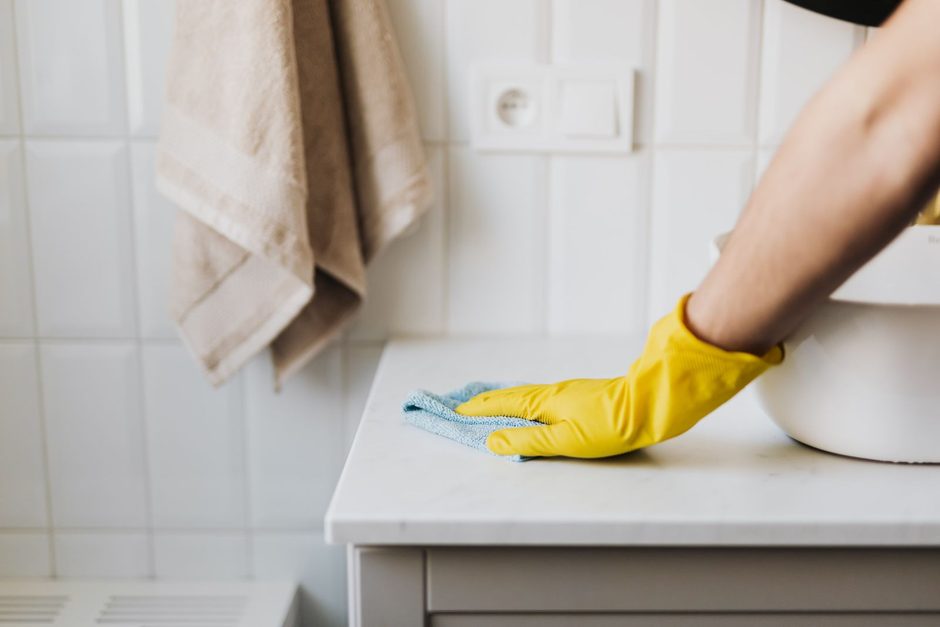 Photo by https://kaboompics.com/ on Pexels Yellow-gloved hand scrubbing a white bathroom basin counter against tiled wall. Photo by https://kaboompics.com/ on Pexels