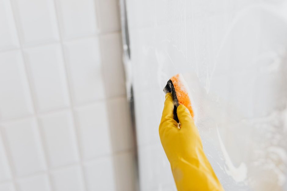 Yellow-gloved hand wiping white bathroom wall tiles with a cleaning cloth. Photo by https://kaboompics.com/ on Pexels