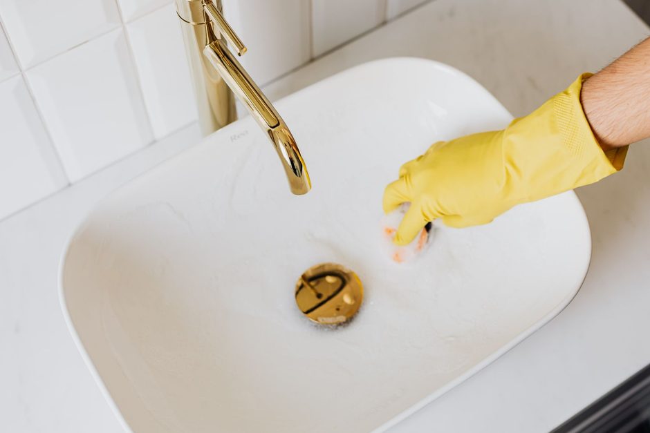 Photo by https://kaboompics.com/ on Pexels Yellow-gloved hand cleaning a white bathroom sink with brass tap. Photo by https://kaboompics.com/ on Pexels