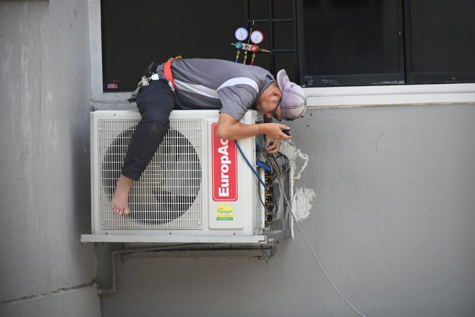 Technician cleaning an air conditioner unit mounted on an apartment building wall. Photo by Richard Low Hong on Pexels